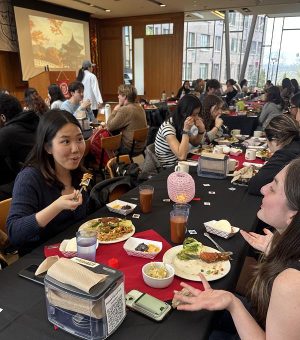 Two students chat with plates of food at a table. Other diners are in the background.