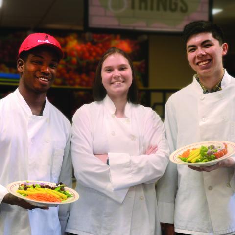Three people in chef jackets, two holding plates of food