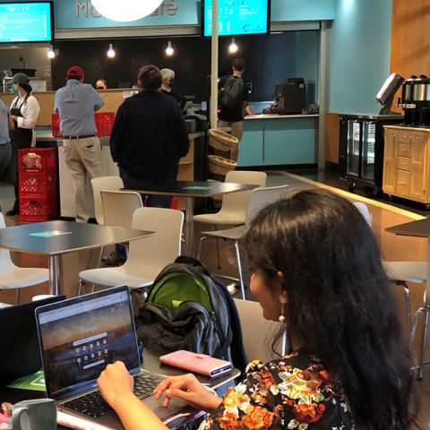 A person sits at a laptop at a cafe table, while in the background people wait at the cafe counter