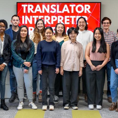Fifteen people stand, posing, in front of a red sign with white text that says, "Translator Interpreter Program."