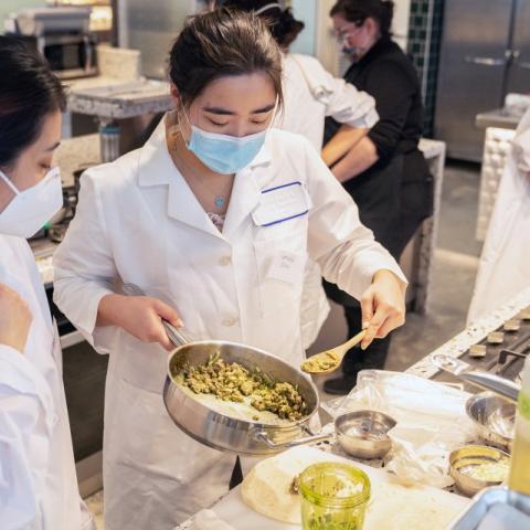 Masked students prepare food in an industrial kitchen.