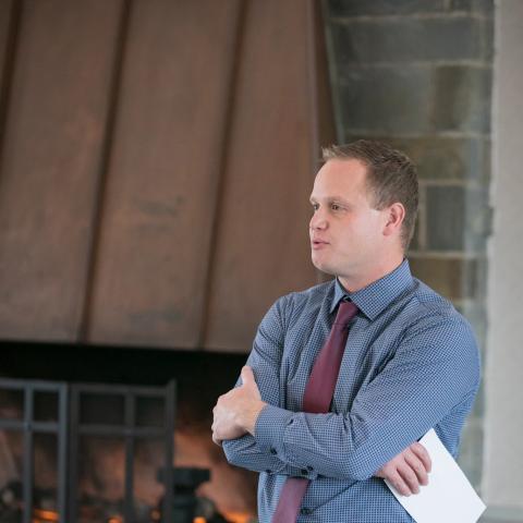 A man wearing a tie speaking with arms folded. A fireplace is in the background.
