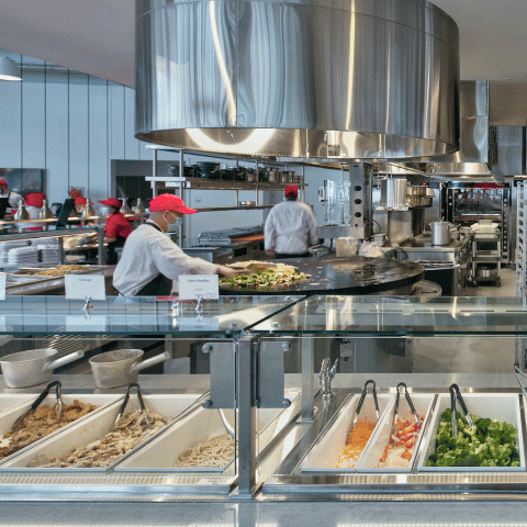 Culinary staff cook on a flat-top grill beyond a row of salad bar style food containers