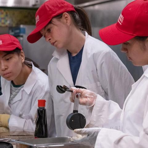 Three students in chef coats and red Cornell Dining caps prepare food in a kitchen