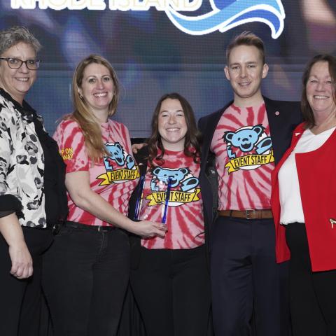 Five people smiling. The three in the middle are wearing tie-dye shirts that say event staff, and there's a lucite award plaque being held by one