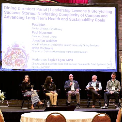 Five people sit in chairs on a stage in front of a projector screen, with one speaking into a microphone