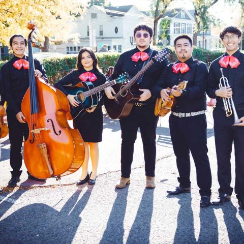 Eight-member mariachi band in traditional black uniforms with red bow ties poses outdoors, holding various instruments including strings and brass, with autumn trees and buildings behind them.