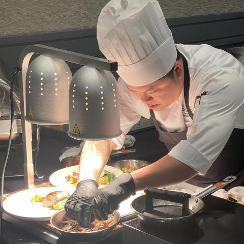 A person wearing a chef jacket and hat and apron bends over a cooking station plating the food