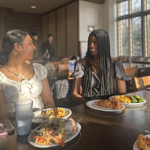 Students smile at each other at a table with plates of food in front of them