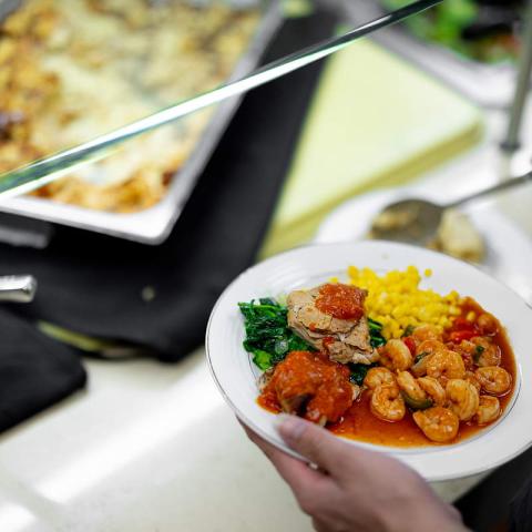A hand holds a plate of food including shrimp in front of serving trays