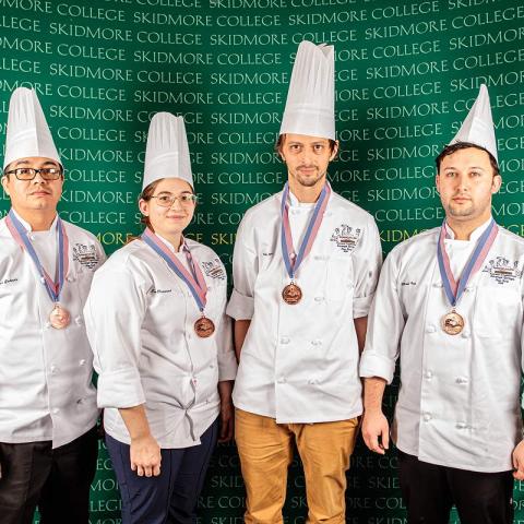 Four people wear chef coats and hats, and medals, in front of a wall that says Skidmore College