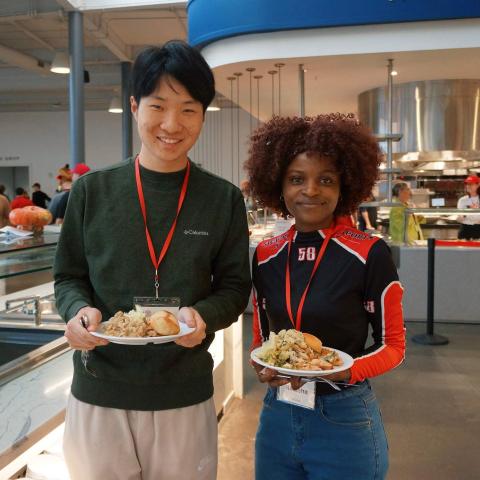 Two smiling people stand in a dining room holding plates of food