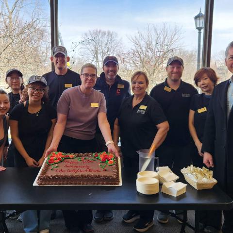 Smiling people wearing cafe uniforms around a table. One person is holding up the edge of a cake.
