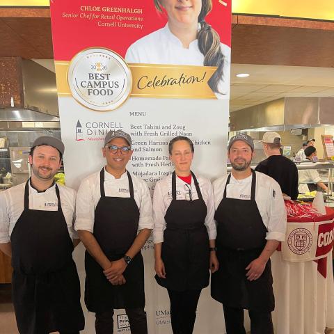 Four people in chef coats and aprons in front of a poster with a woman's face