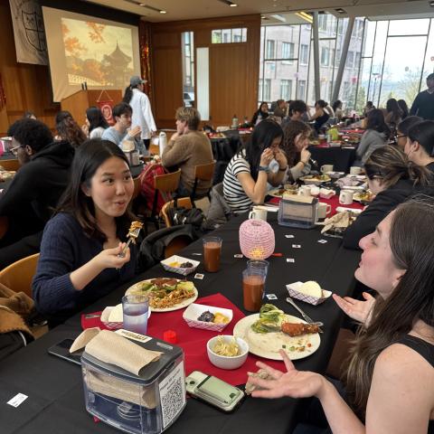 Two students chat with plates of food at a table. Other diners are in the background.
