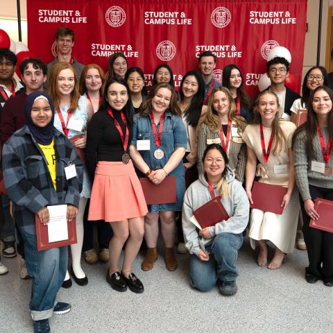 Cornell students wearing medals and holding certificates pose with staff in front of a red Student & Campus Life backdrop.