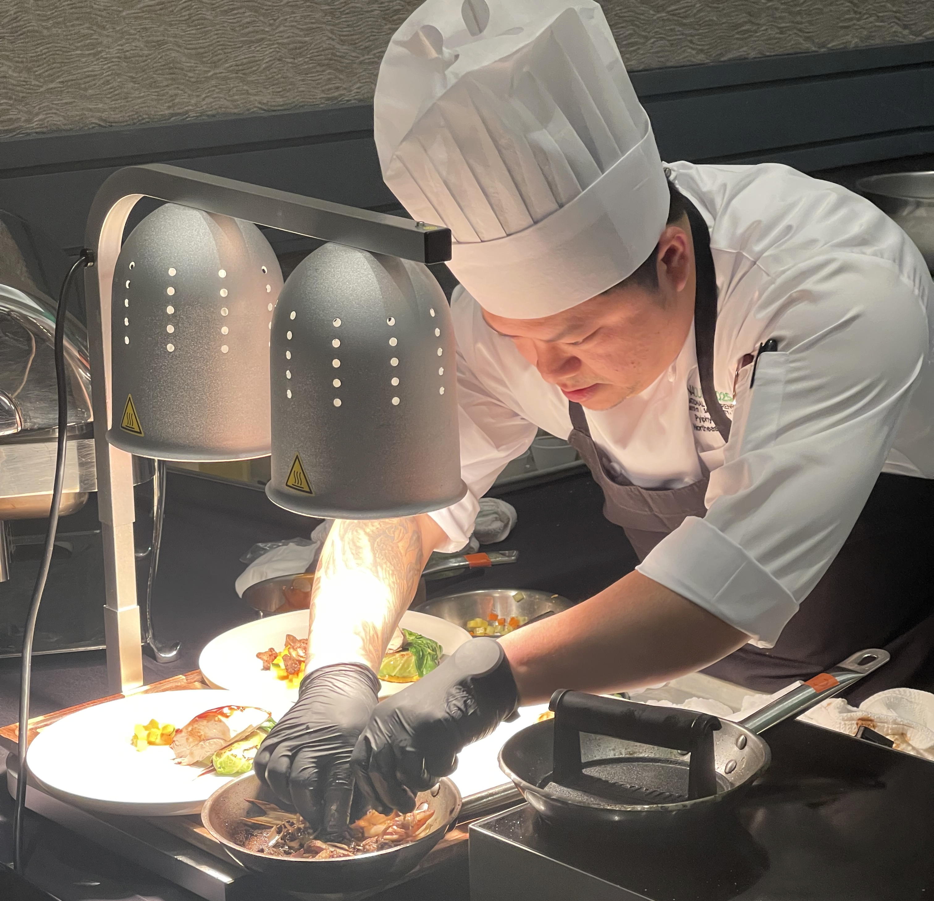 A person wearing a chef jacket and hat and apron bends over a cooking station plating the food