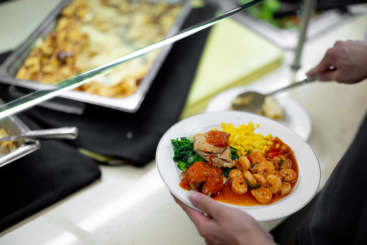 A hand holds a plate of food including shrimp in front of serving trays