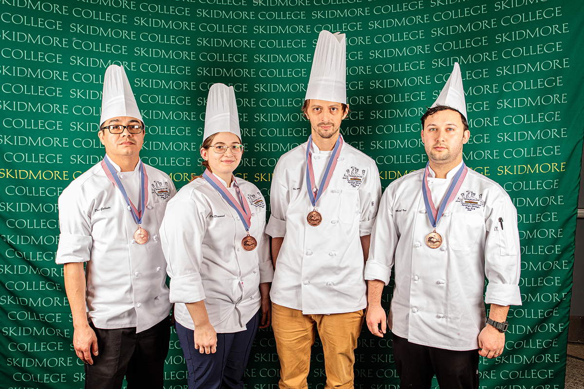 Four people wear chef coats and hats, and medals, in front of a wall that says Skidmore College