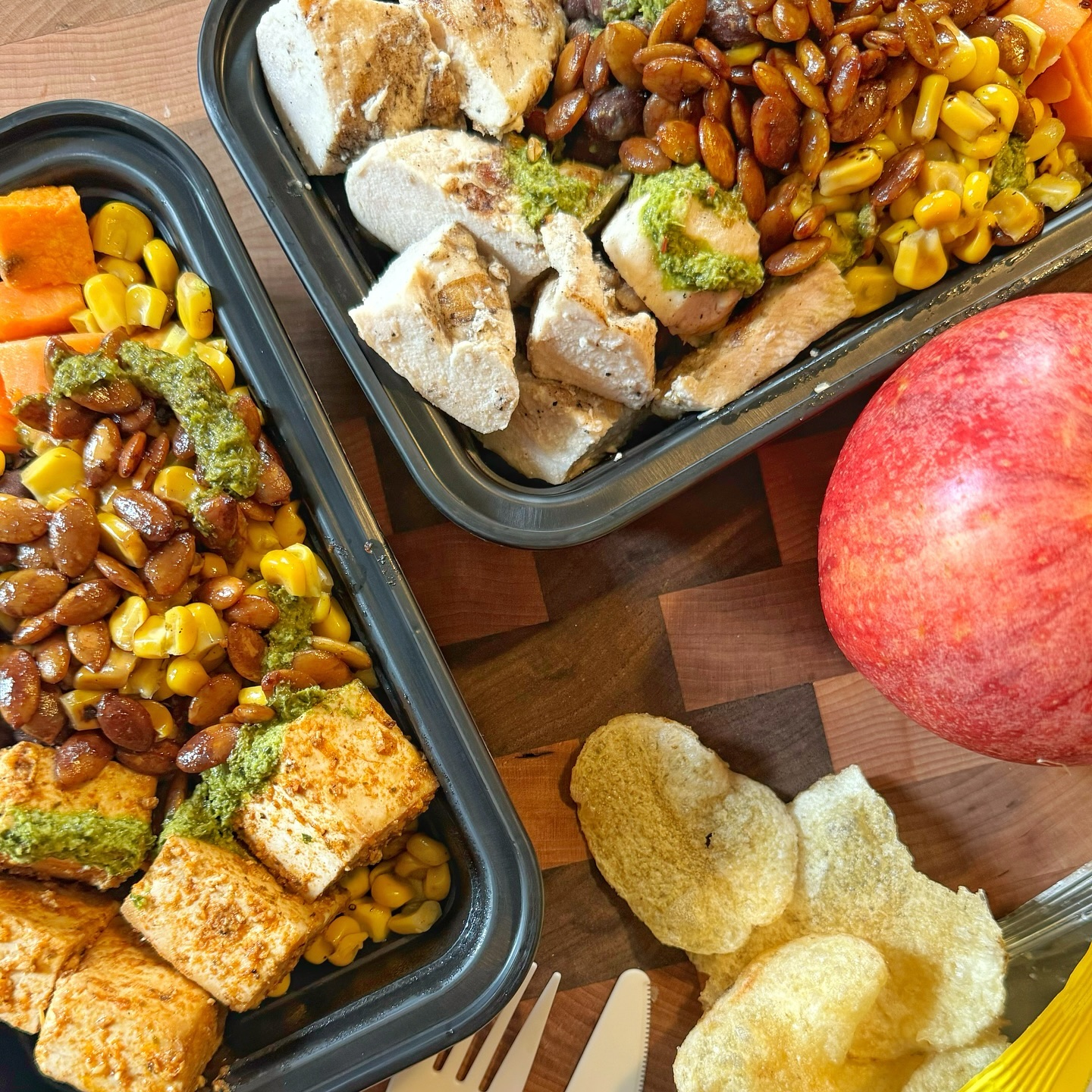 Two takeout containers full of food, with potato chips and an apple on the table next to them