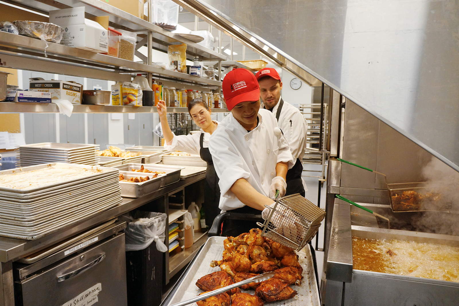 A person in a kitchen takes fried chicken out of a fryer basket, with two others looking on