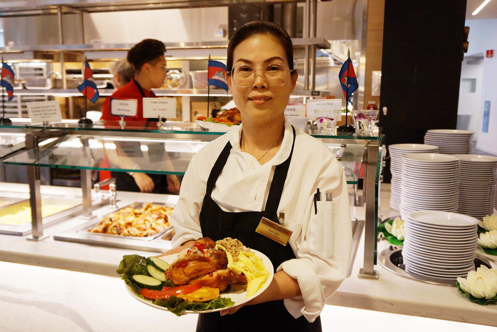 A person in an apron holds a plate of food in front of a serving line