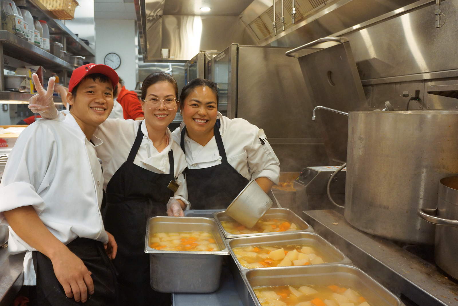 Three people wearing chef coats and aprons in a kitchen with pots of soup