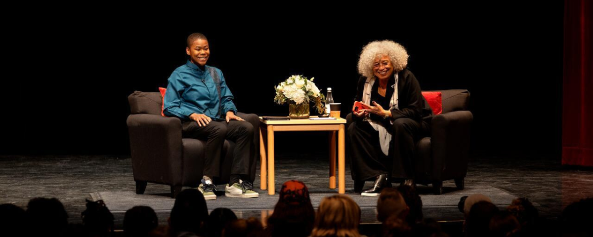 Angela Davis sits on stage with a student at a packed event.