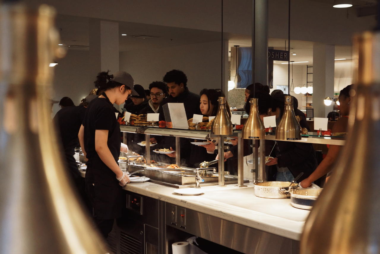 A staff member serves food with a line of students on the other side of the counter