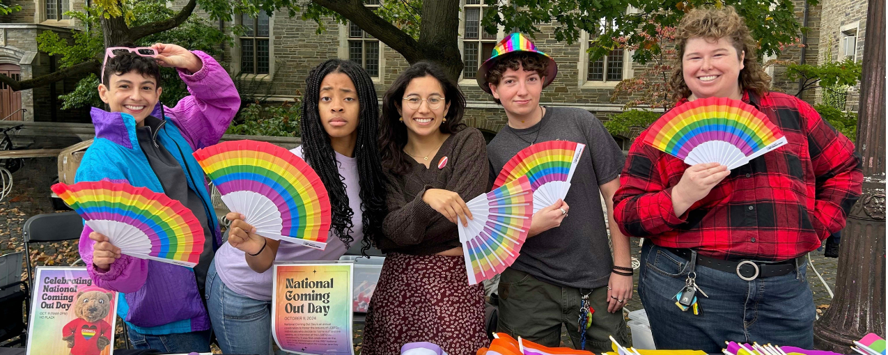 Students pose while tabling for National Coming Out Day. Students hold rainbow fans.
