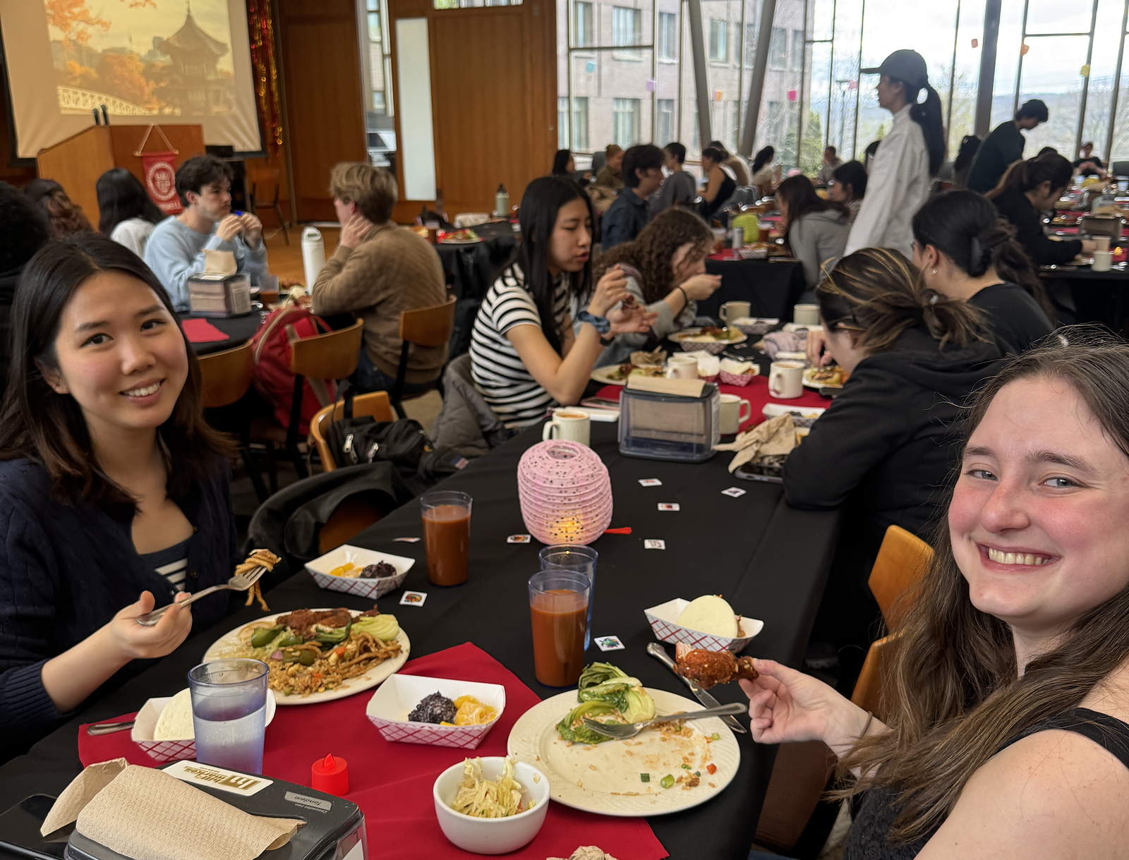 Smiling students with plates of food at a table, with more diners in the background