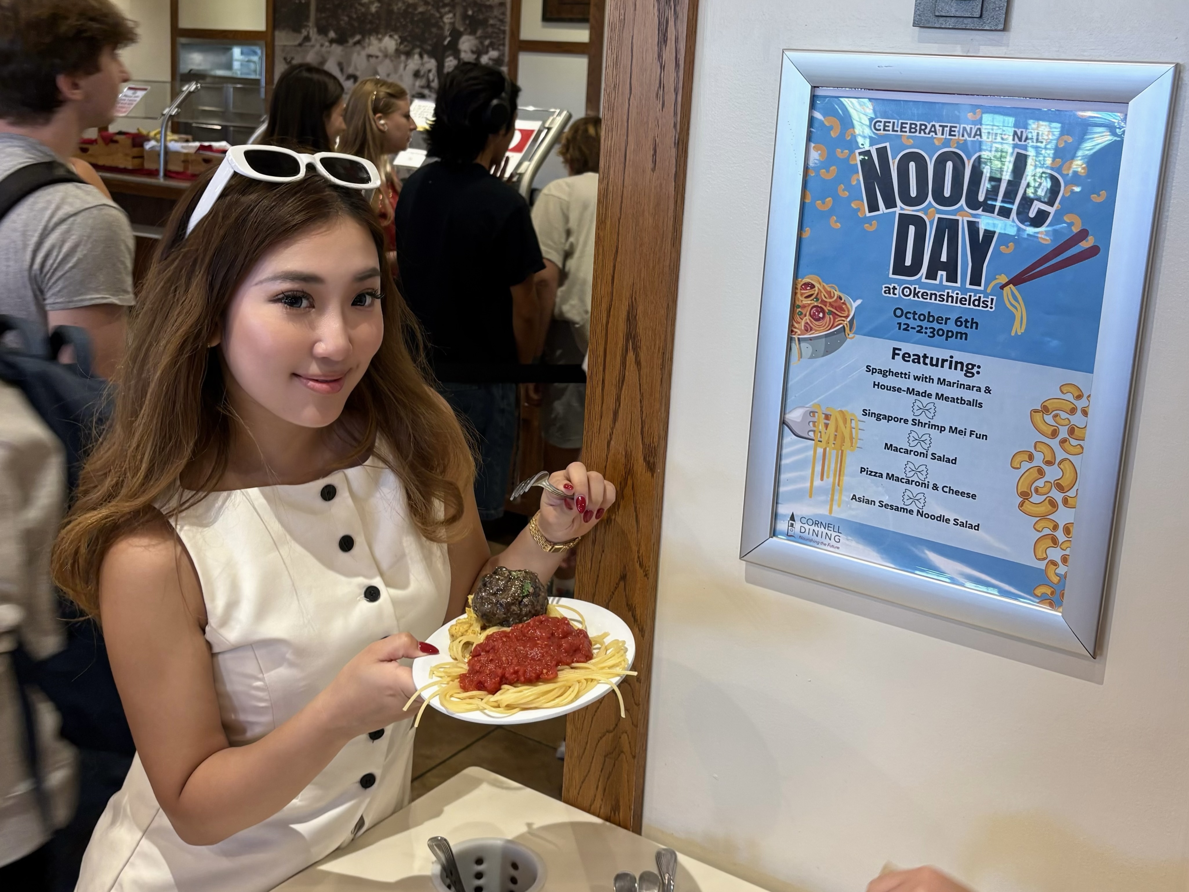 A smiling person with glasses on top of her head holds a plate of spaghetti and sauce. A poster says celebrate National Noodle Day.
