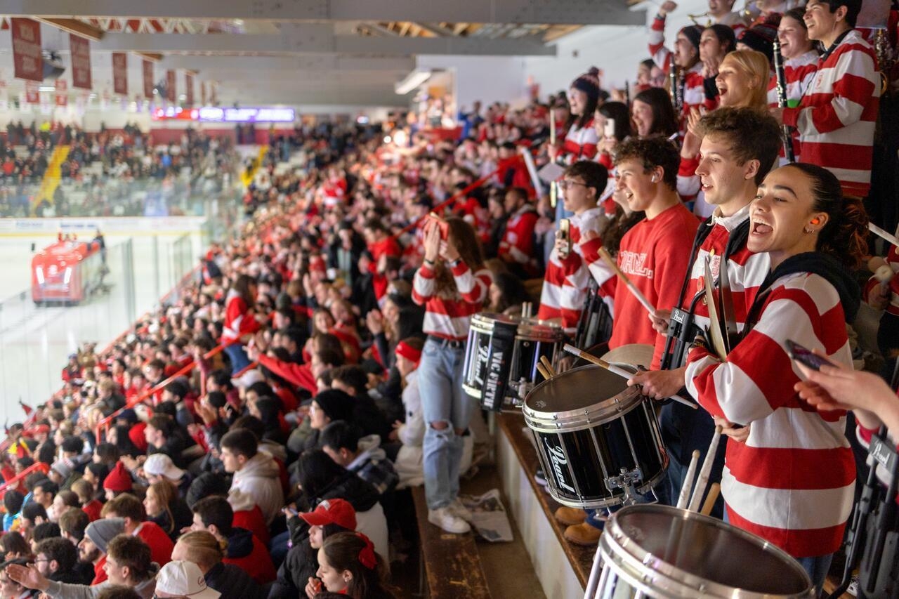 Cornell students in red-and-white jerseys play drums and cheer alongside a packed crowd at an ice hockey game