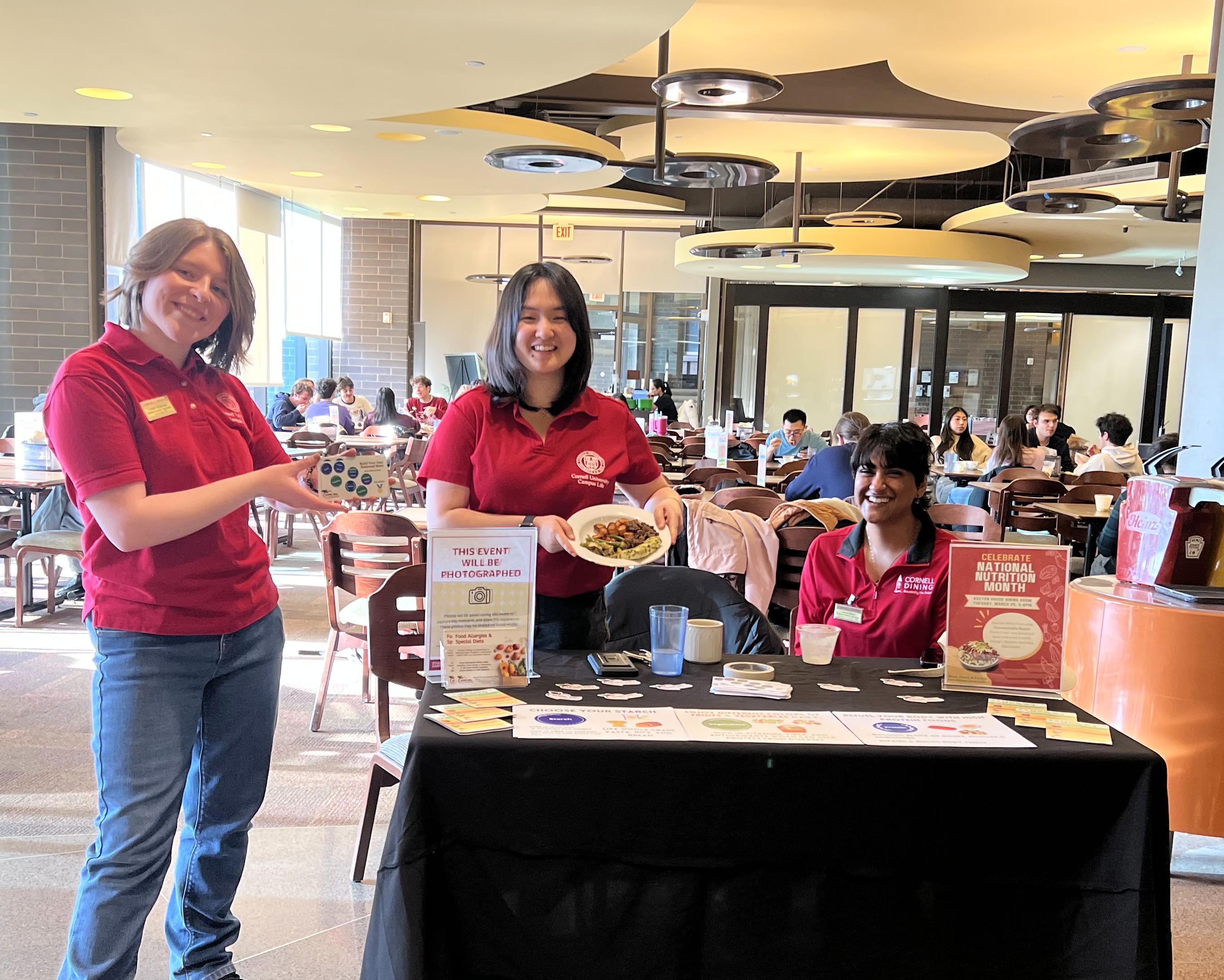Three people wearing red Cornell shirts at a table with info and food samples. People are at tables eating in the background.