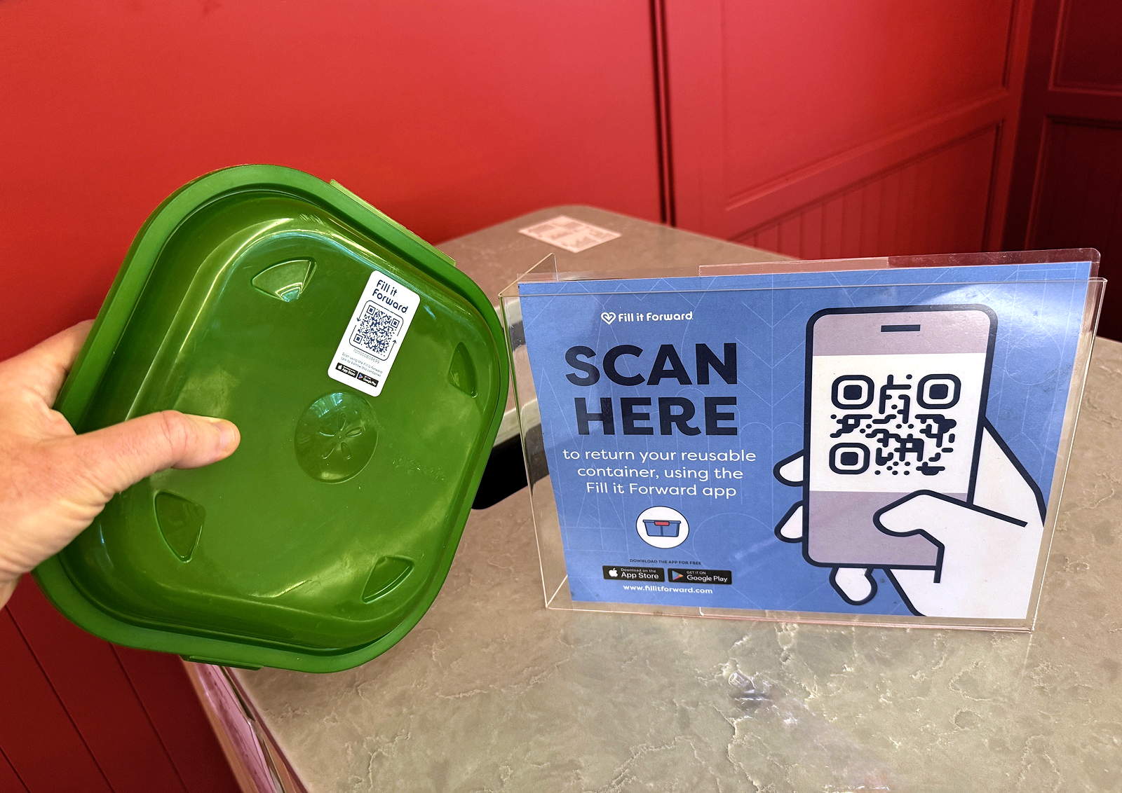A hand holds a green plastic food takeout container next to a sign that says scan here to return your container