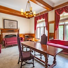A common area with a long wooden table with two chairs, two couches, a lounge chair, a fireplace and two large windows with curtains