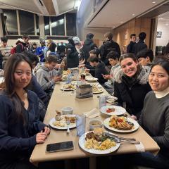 A group sits around a dining room table with plates of food in front of them. Others are standing beyond the table.