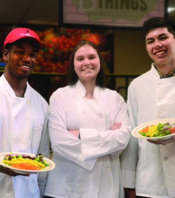 Three people in chef jackets, two holding plates of food