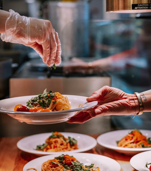 A hand sprinkles herbs over a plate of pasta being held by a diner