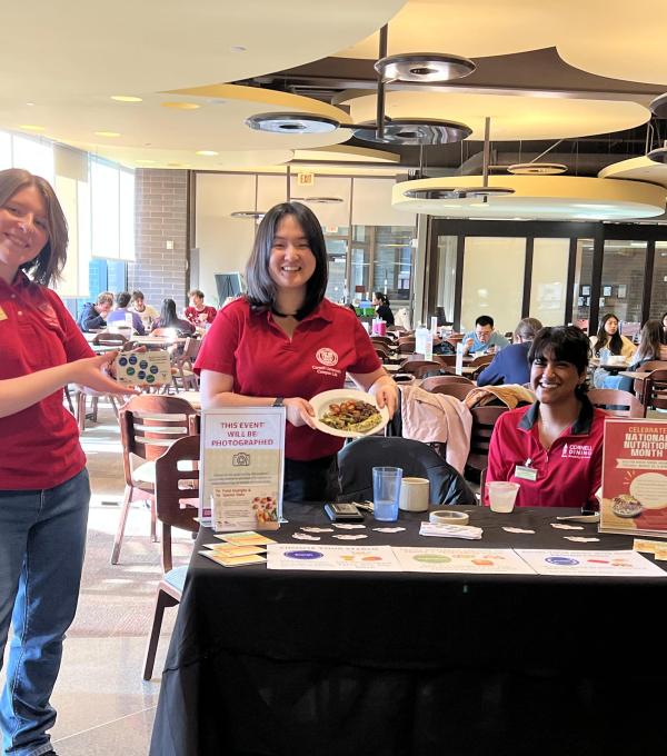 Three people in red Cornell shirts at a table with samples and posters in a dining room, with people eating at tables in the background