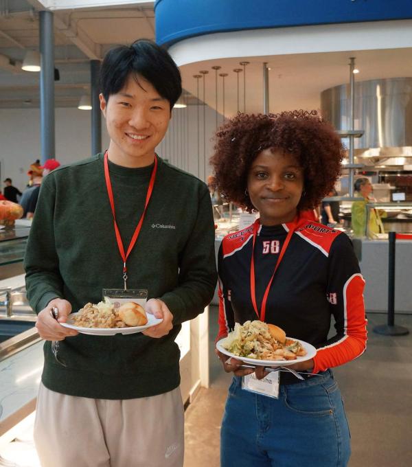 Two smiling people stand in a dining room holding plates of food