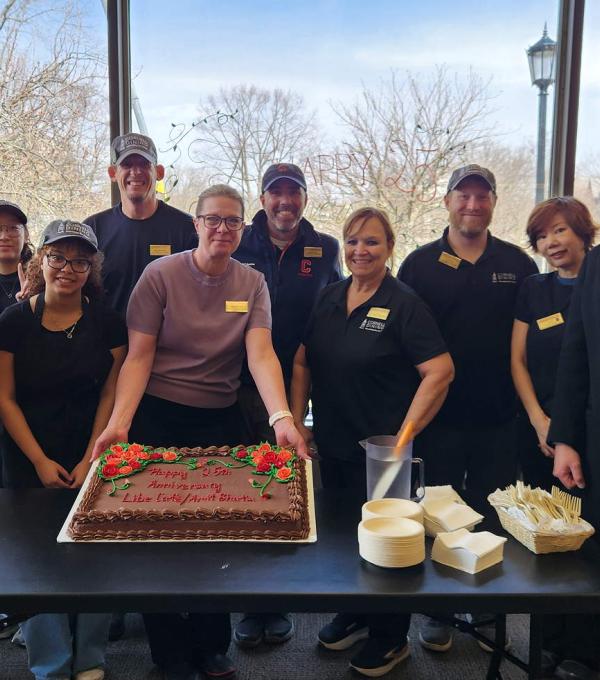 Smiling people wearing cafe uniforms around a table. One person is holding up the edge of a cake.