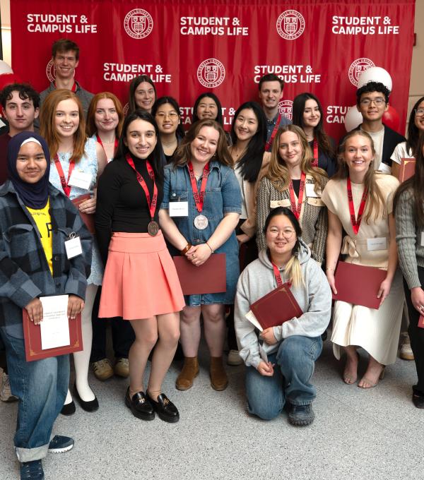 Cornell students wearing medals and holding certificates pose with staff in front of a red Student & Campus Life backdrop.