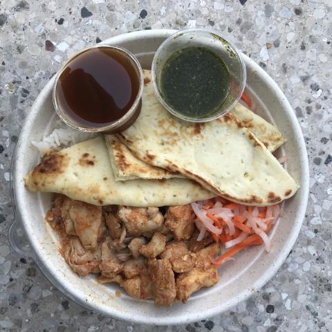 Bowl of rice topped with chicken and vegetables, with a piece of naan and two sauce containers