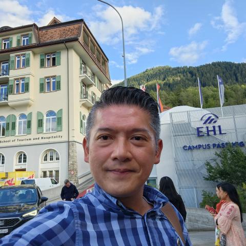 A person poses for a selfie in front of a school building with a tree-covered hillside beyond