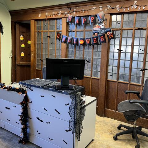 A desk and rolling office chair in front of a wood and frosted glass wall. Halloween decorations include a Happy Halloween banner on the wall, and a spiderweb drape and paper bats on the desk
