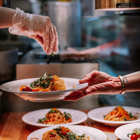 A hand sprinkles herbs over a plate of pasta being held by a diner