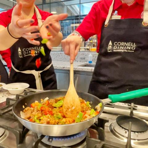 People wearing Cornell Dining aprons stand at a stovetop and put food into a pan and stir it
