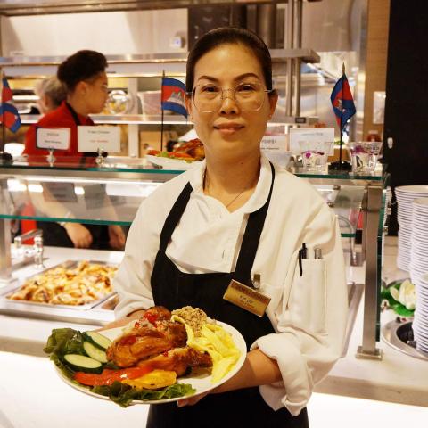 A person in an apron holds a plate of food in front of a serving line