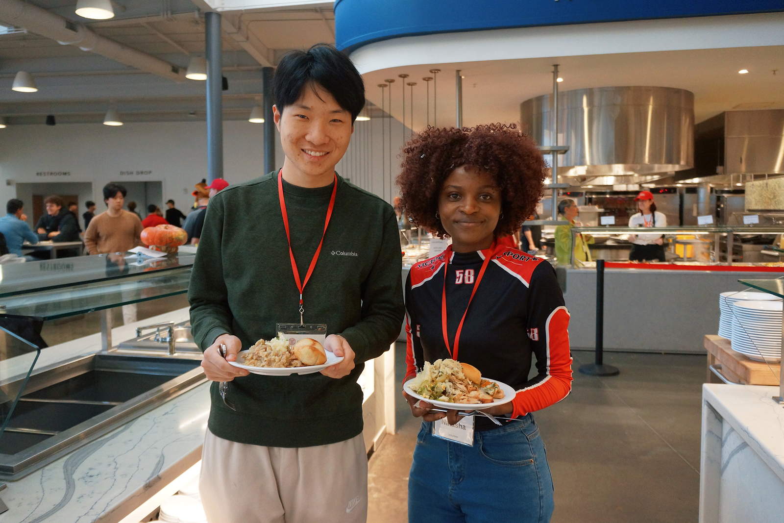 Two smiling people stand in a dining room holding plates of food